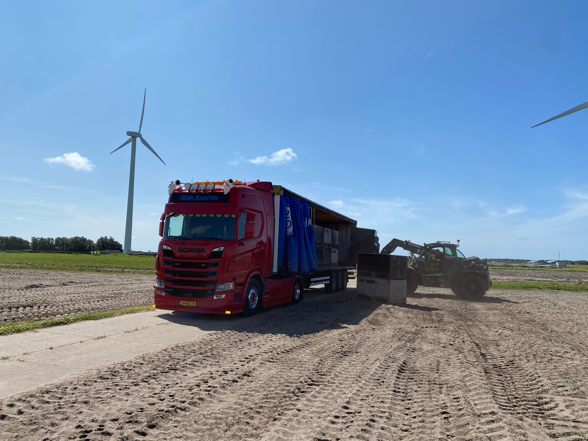 gebr-karsten-transport rode vrachtwagen wordt geladen door tractor in veld met blauwe lucht en windturbines