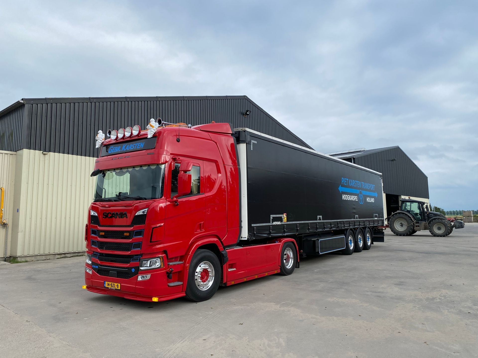 Rode vrachtwagen met zwarte oplegger geparkeerd bij een donker gebouw en een tractor op een bewolkte dag.