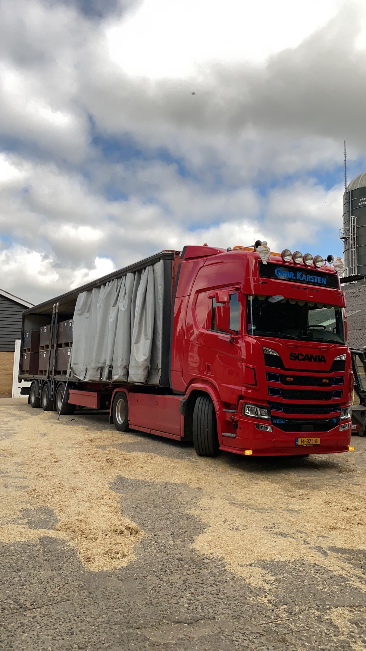 Rode vrachtwagen met overdekte aanhanger geparkeerd op een grindterrein onder een bewolkte hemel.