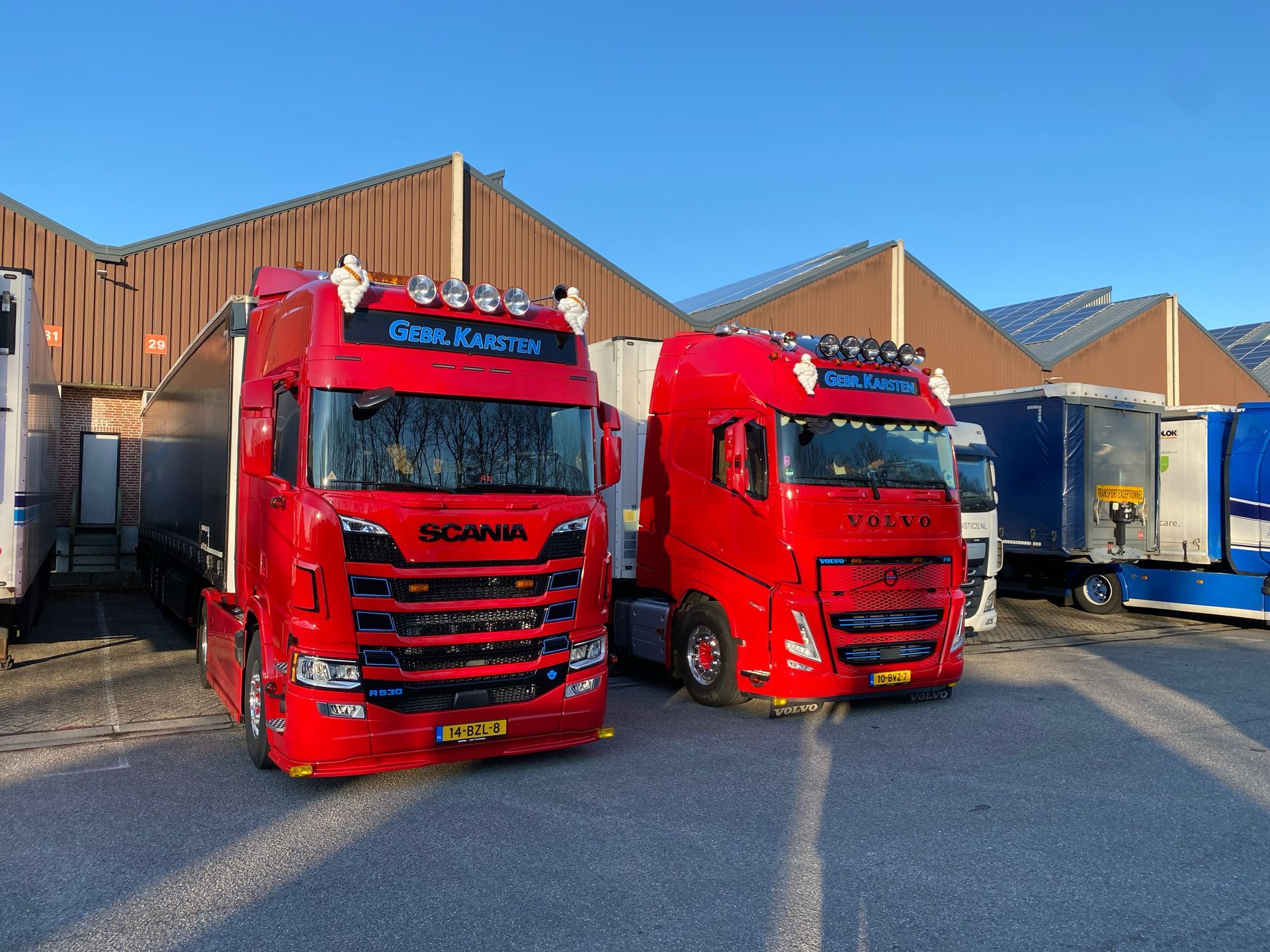 Twee rode vrachtwagens geparkeerd buiten, tegen een gebouw, op een zonnige dag.