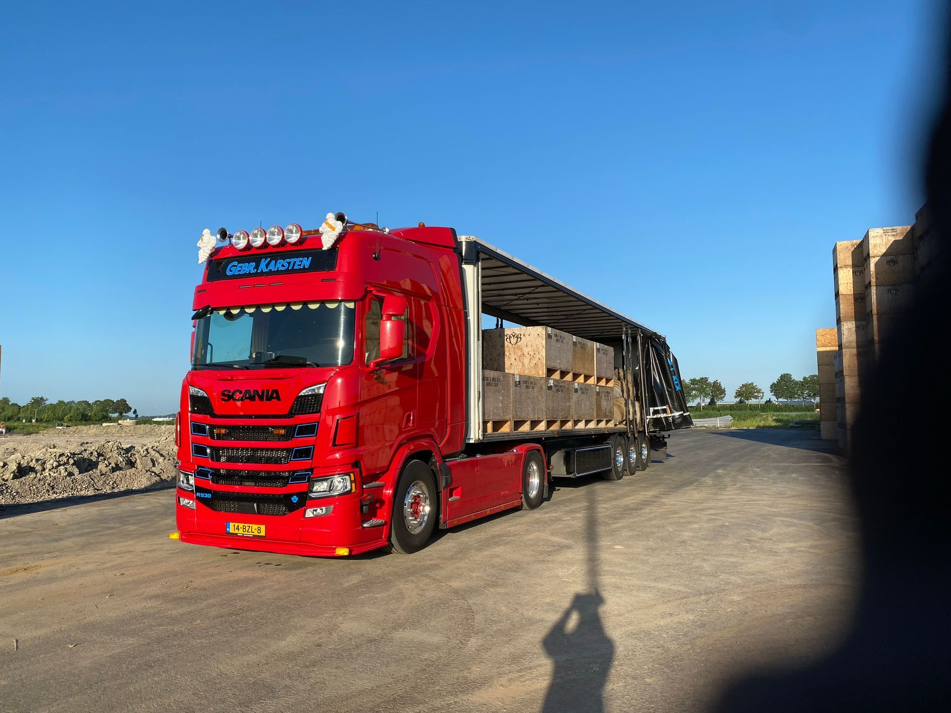 Rode Scania vrachtwagen met oplegger geladen met pallets op een verharde weg onder een blauwe lucht.