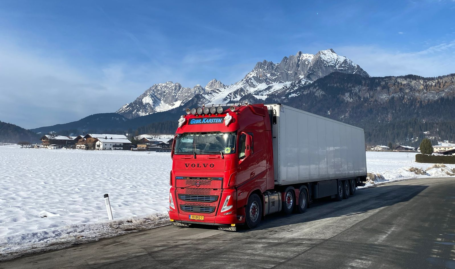 Rode vrachtwagen op een besneeuwde weg met bergen op de achtergrond en een blauwe lucht.