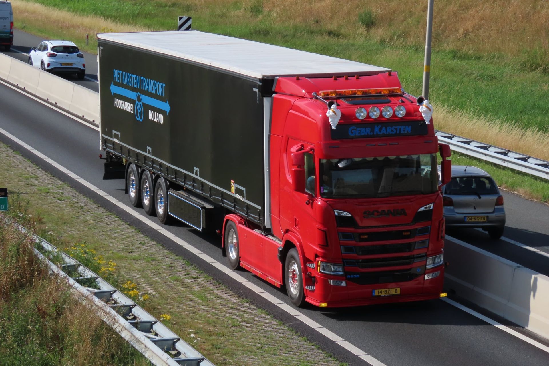 Rode vrachtwagen met zwarte aanhanger op de snelweg. Witte auto op de linkerbaan, groen gebladerte op de achtergrond.