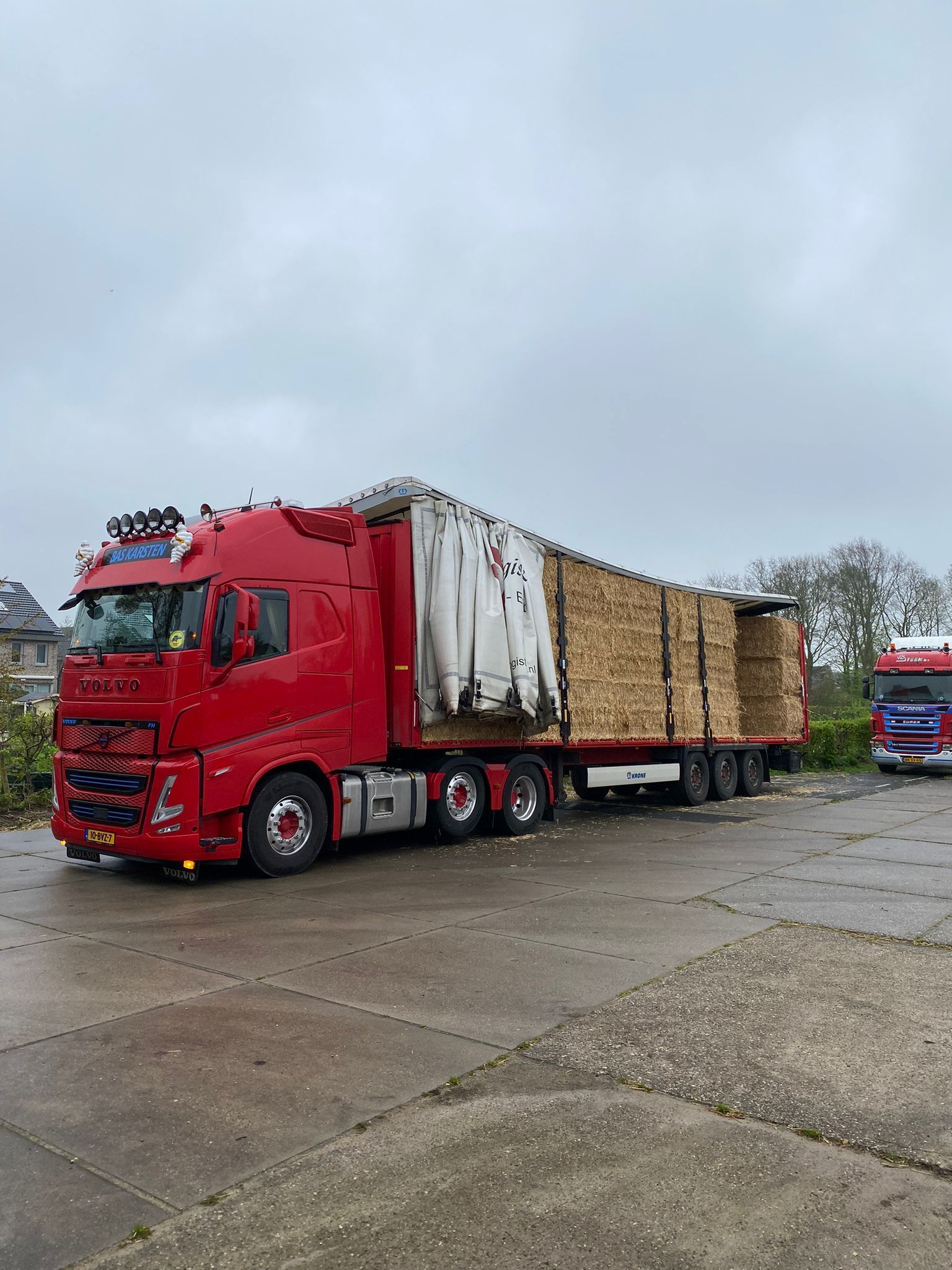 Rode vrachtwagen met opgestapeld hout op een aanhanger, geparkeerd op een betonnen ondergrond onder een bewolkte hemel.