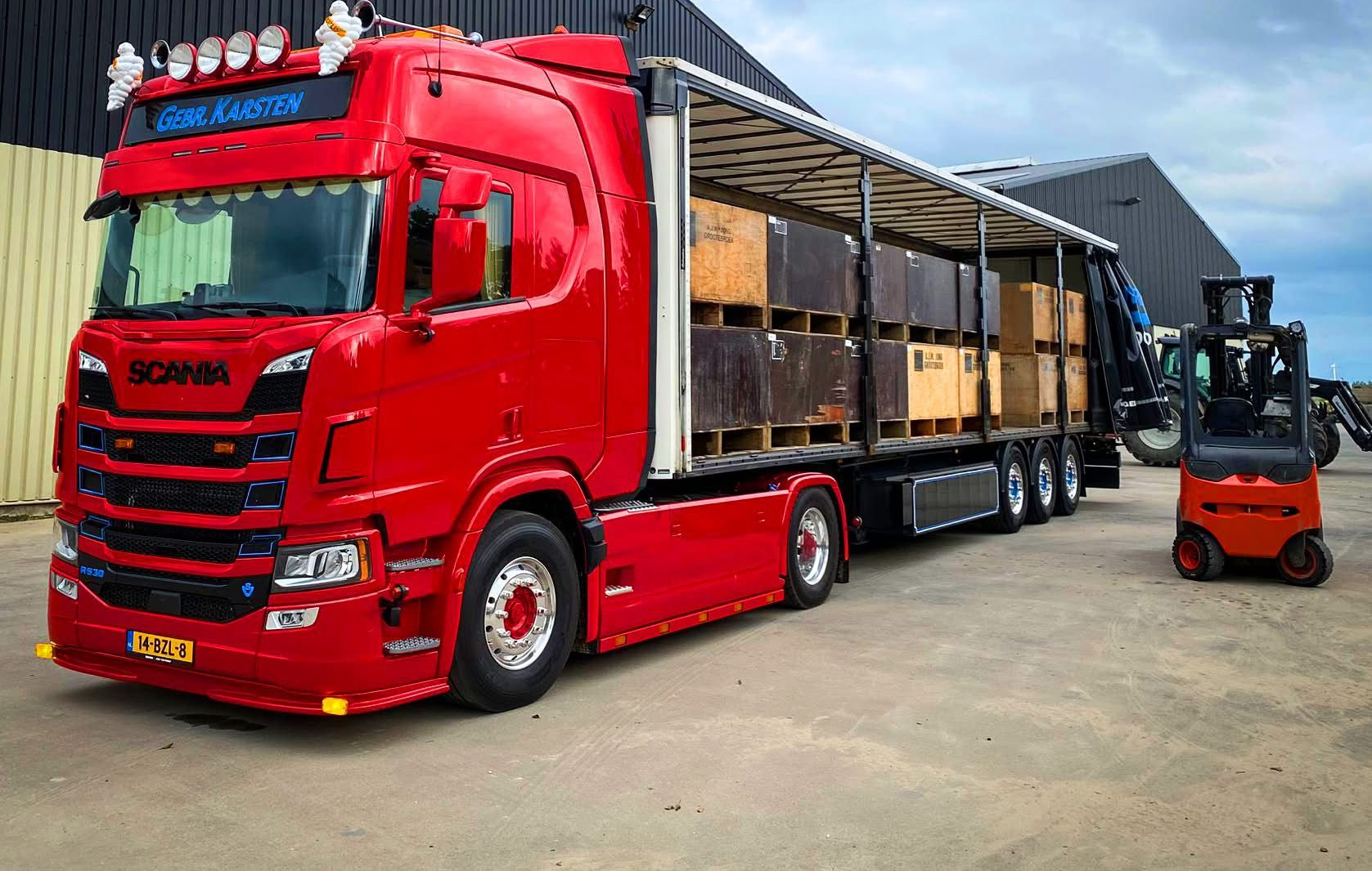 Een rode Scania vrachtwagen geladen met houten kratten; ernaast staat een vorkheftruck klaar om te lossen.