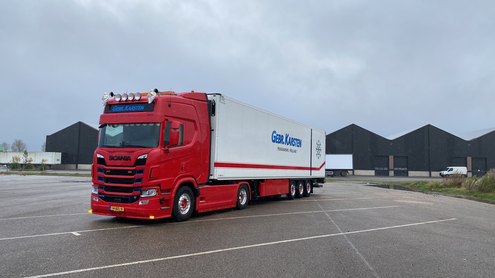 Rood-witte vrachtwagen met aanhanger op een verhard terrein voor donkere gebouwen. Bewolkte lucht.