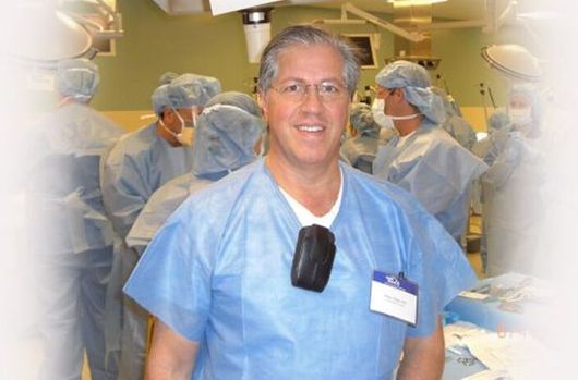 Doctor in blue scrubs smiles in an operating room with a medical team behind him.