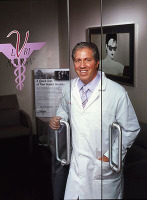 Man in lab coat, at a clinic door, hand on handle; logo on glass, art on wall.