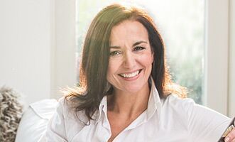 Woman with dark hair, smiling broadly, wearing a white shirt; bright natural light.