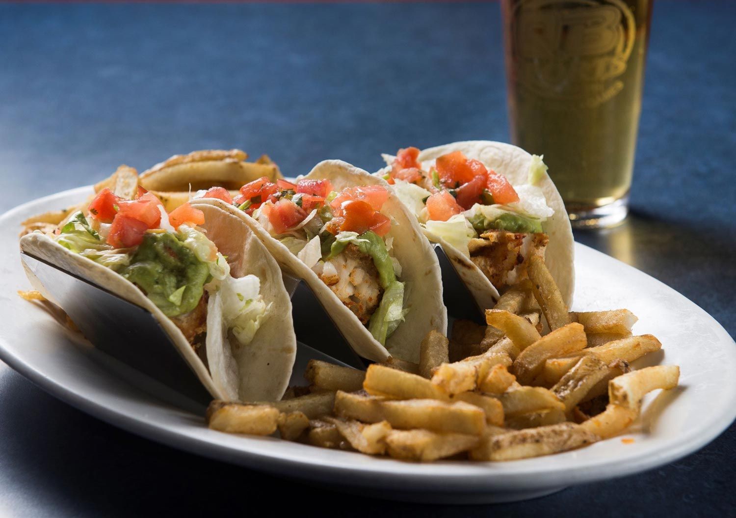 A plate of tacos and french fries next to a glass of beer.
