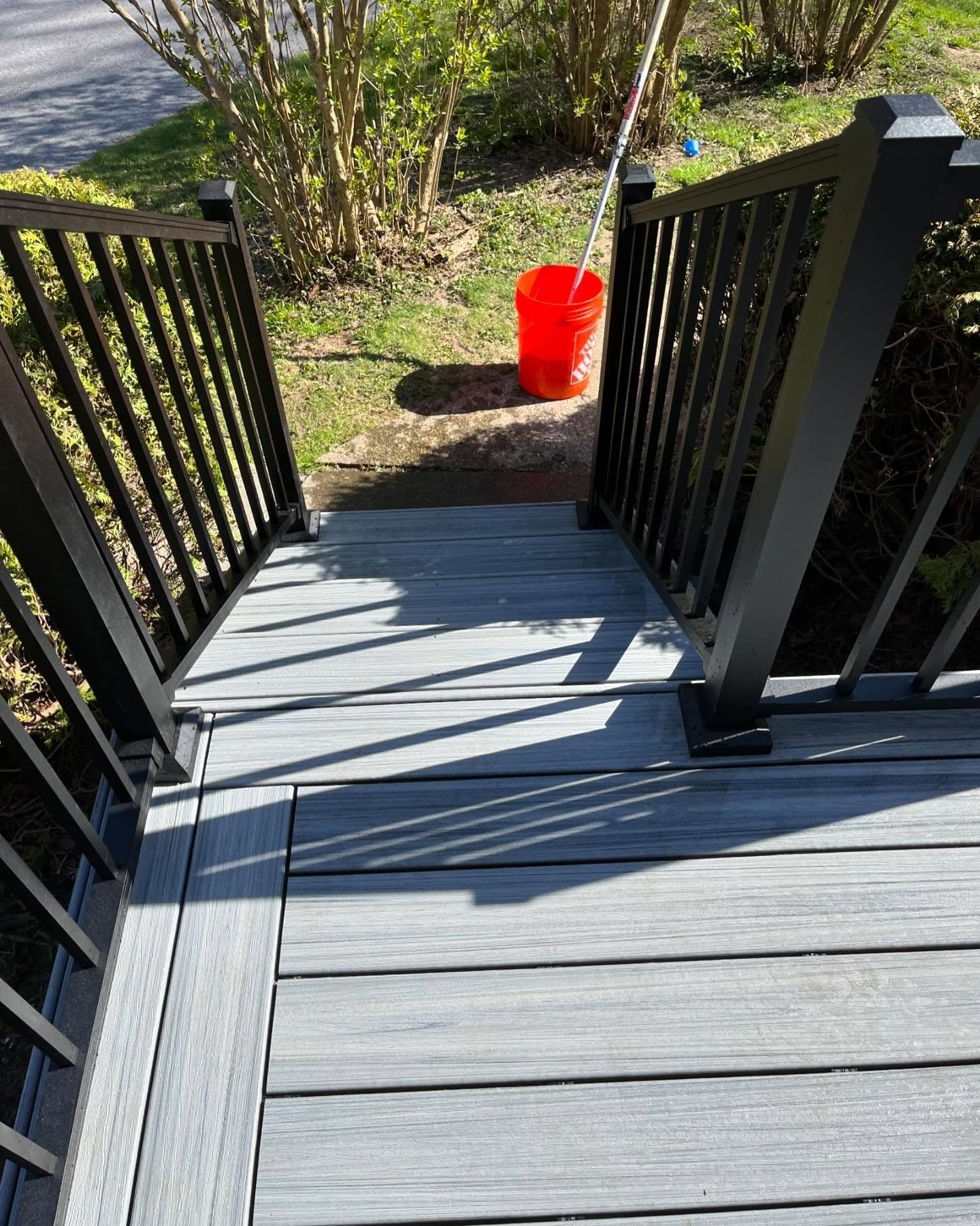Gray deck with black railings and stairs leading to a yard, red bucket in the background.