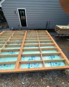 Wooden deck frame under construction, with insulation between supports, gravel ground, and a gray building in the background.