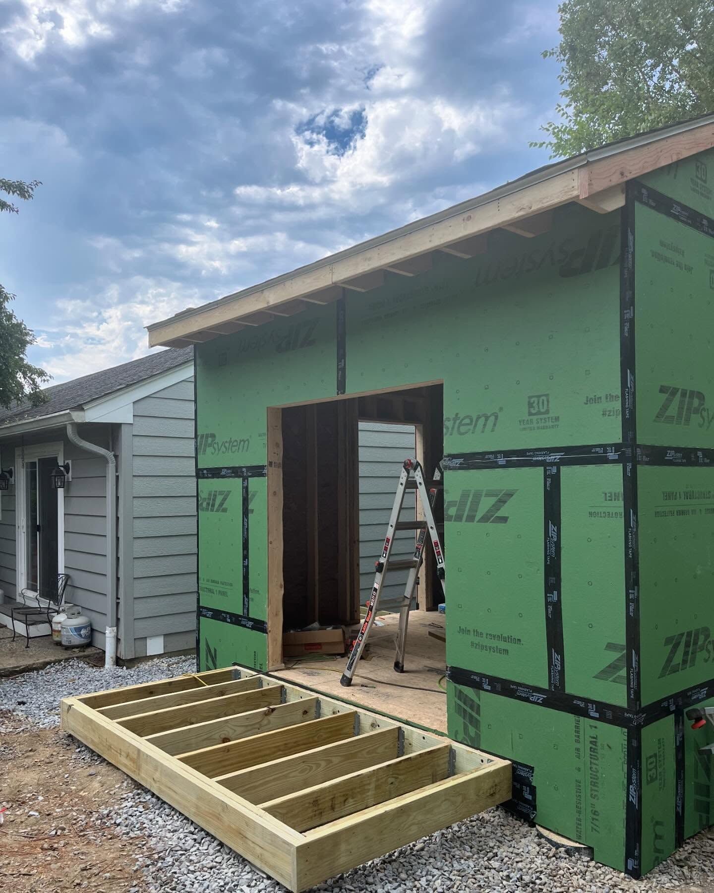 Construction site: a new room addition with green sheathing, attached to a gray house; wooden deck and ladder present.