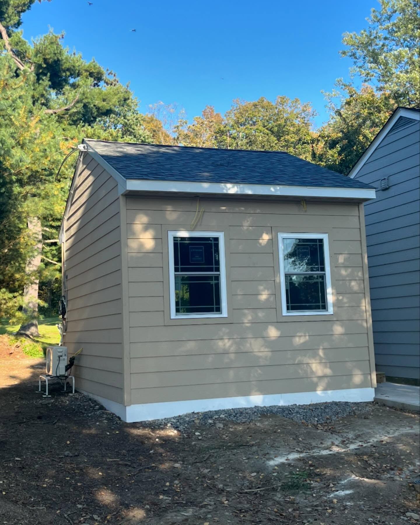 Tan shed with two windows and a dark roof, against a backdrop of trees and a clear sky.