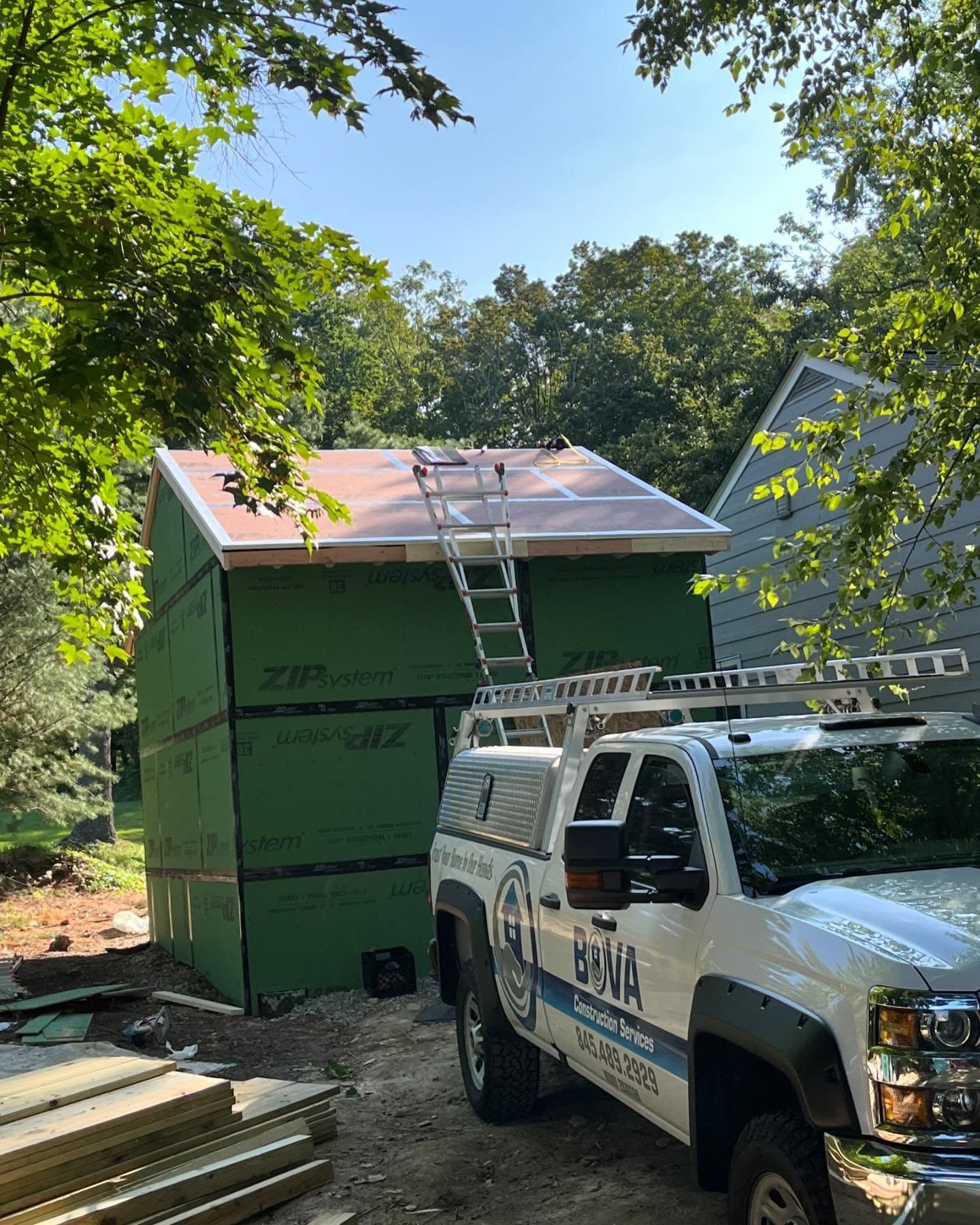 Construction of a shed with a ladder leaning against it, green siding, and a white truck in a wooded area.