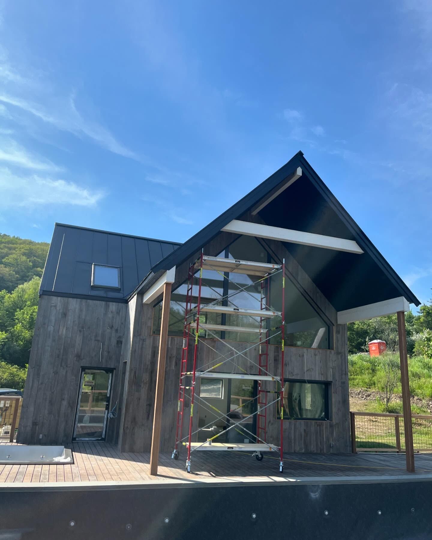 Modern wooden house under construction; scaffolding, glass windows, black roof, blue sky.