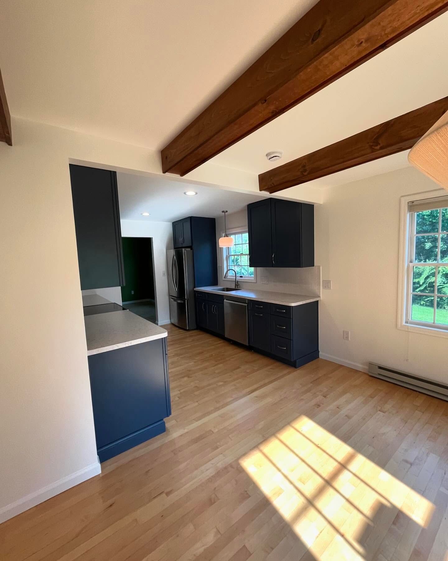Blue kitchen cabinets and white counters in a bright, open space with wooden beams and light wood floors.