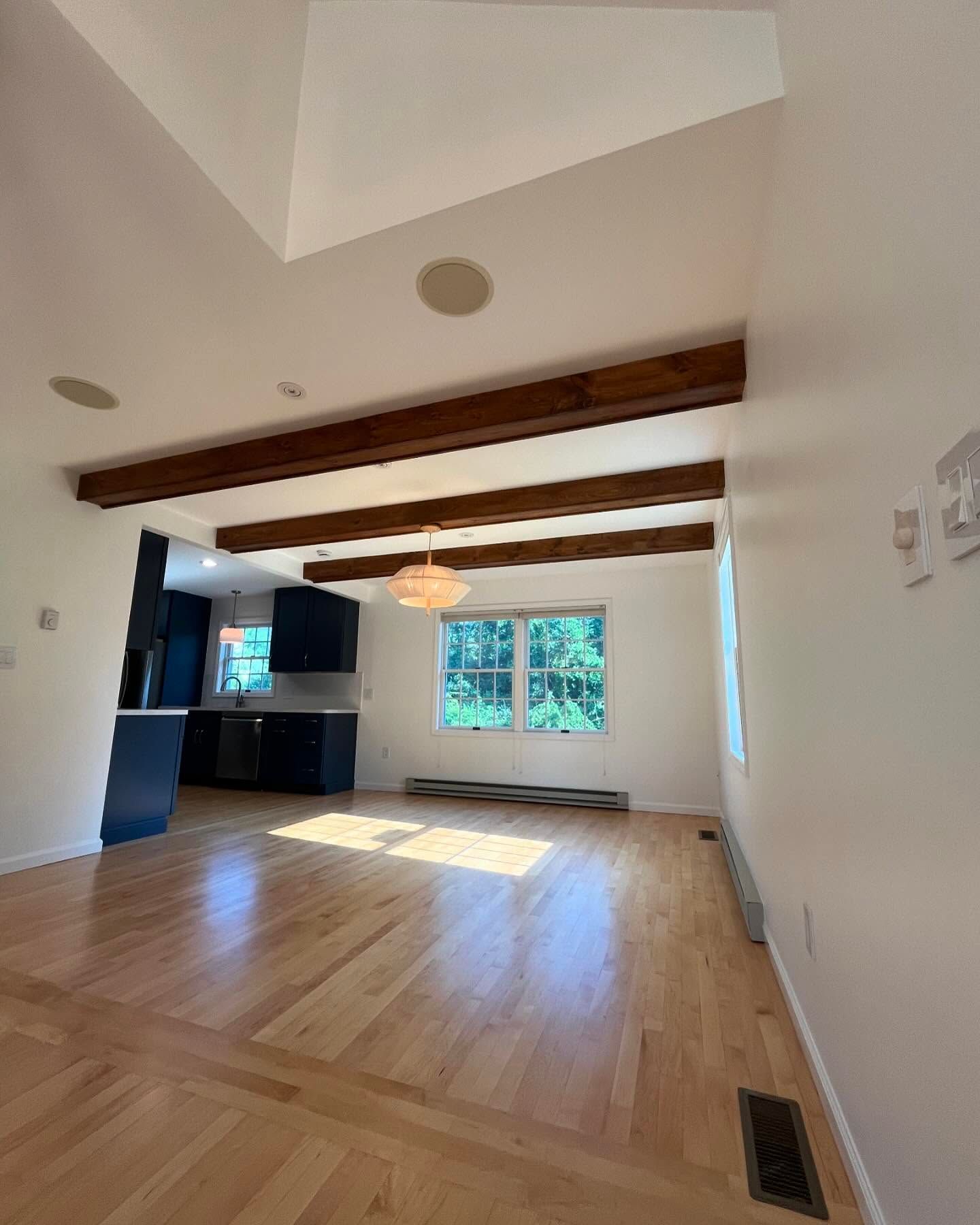 Bright living room with wood beams, hardwood floor, and open kitchen with dark cabinets.
