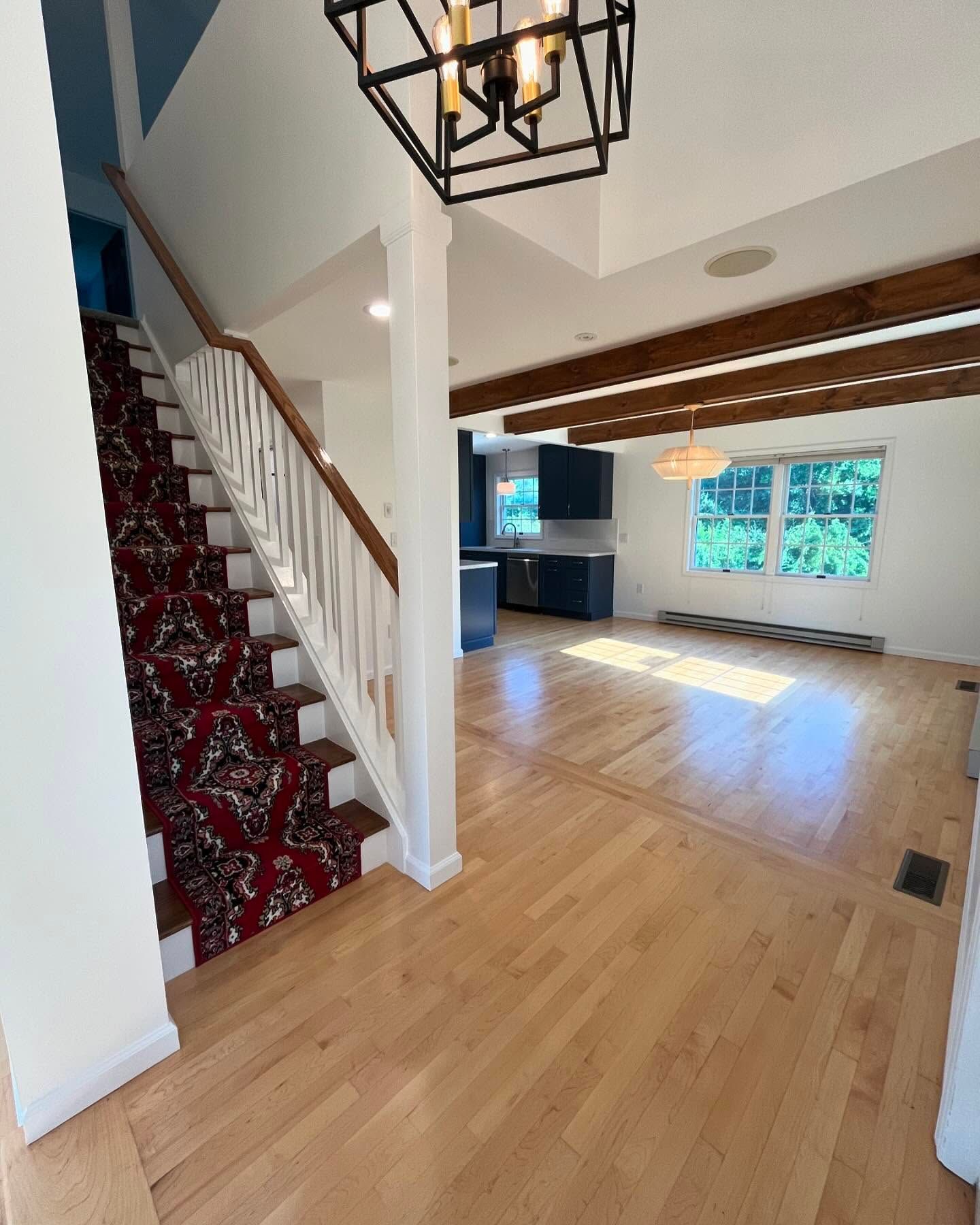 Staircase with patterned runner leads to a light-filled room with beams, hardwood floors, and a kitchen.