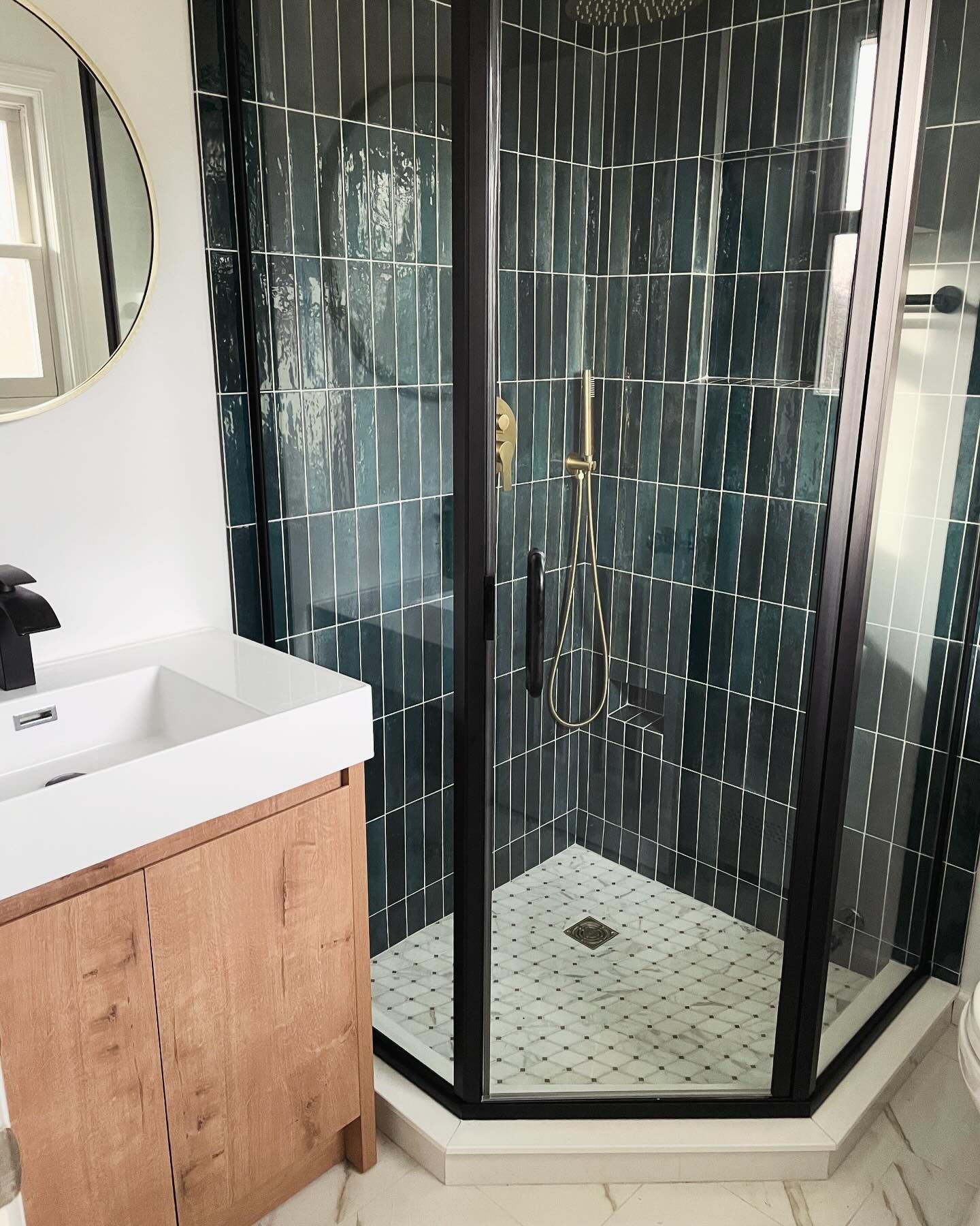 Bathroom with teal vertical tile shower, wood vanity, gold fixtures, and round mirror.