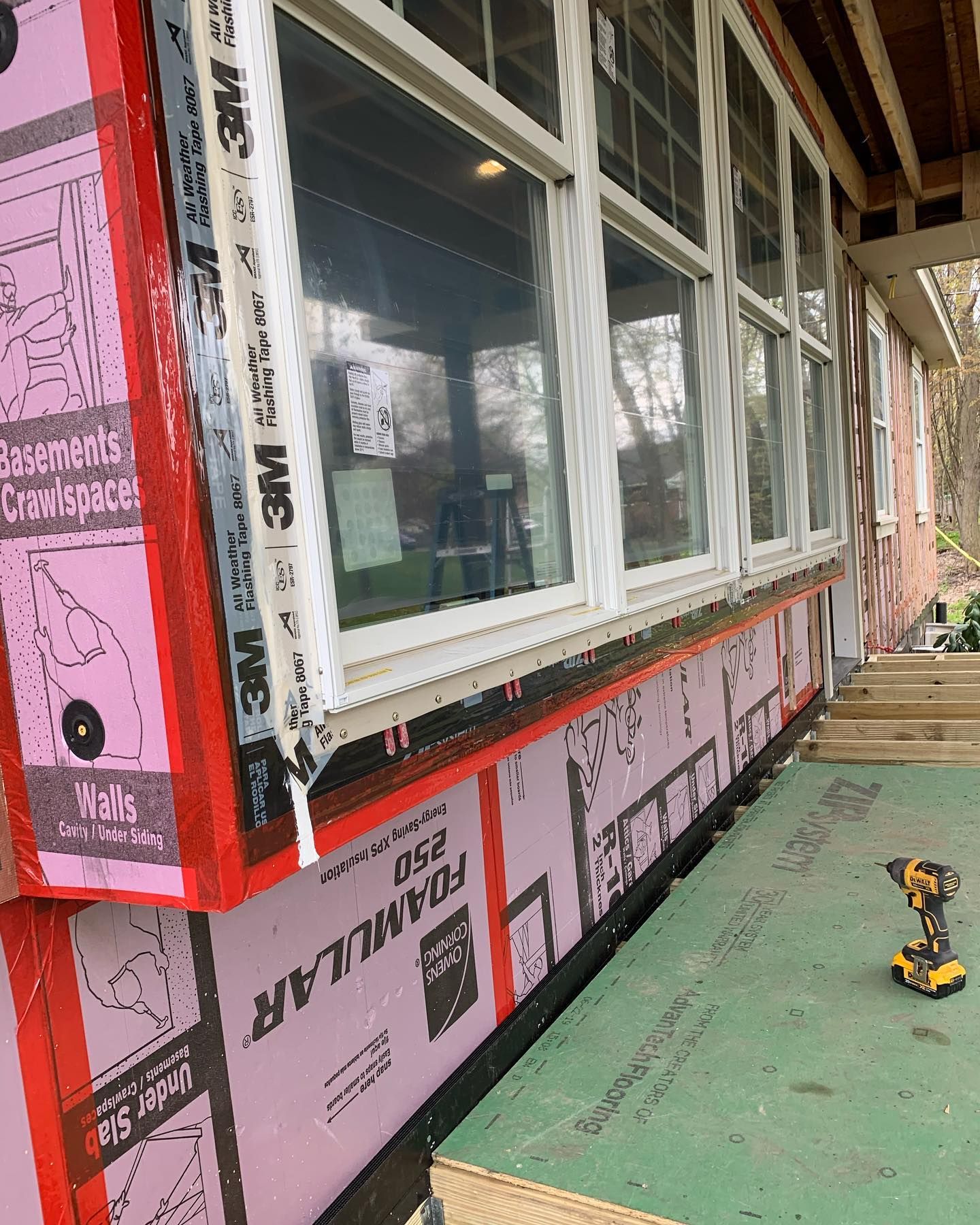 Exterior of a house under construction. Pink foam insulation installed below windows with a drill on a green board.