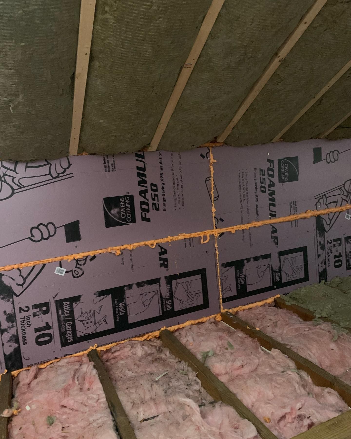 Attic with pink fiberglass insulation, purple foam board, and exposed wooden beams.