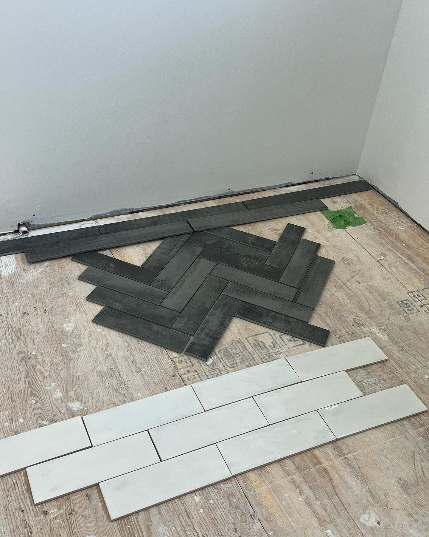 Herringbone tile pattern in progress; dark gray and white tiles on a wood floor near a gray wall.