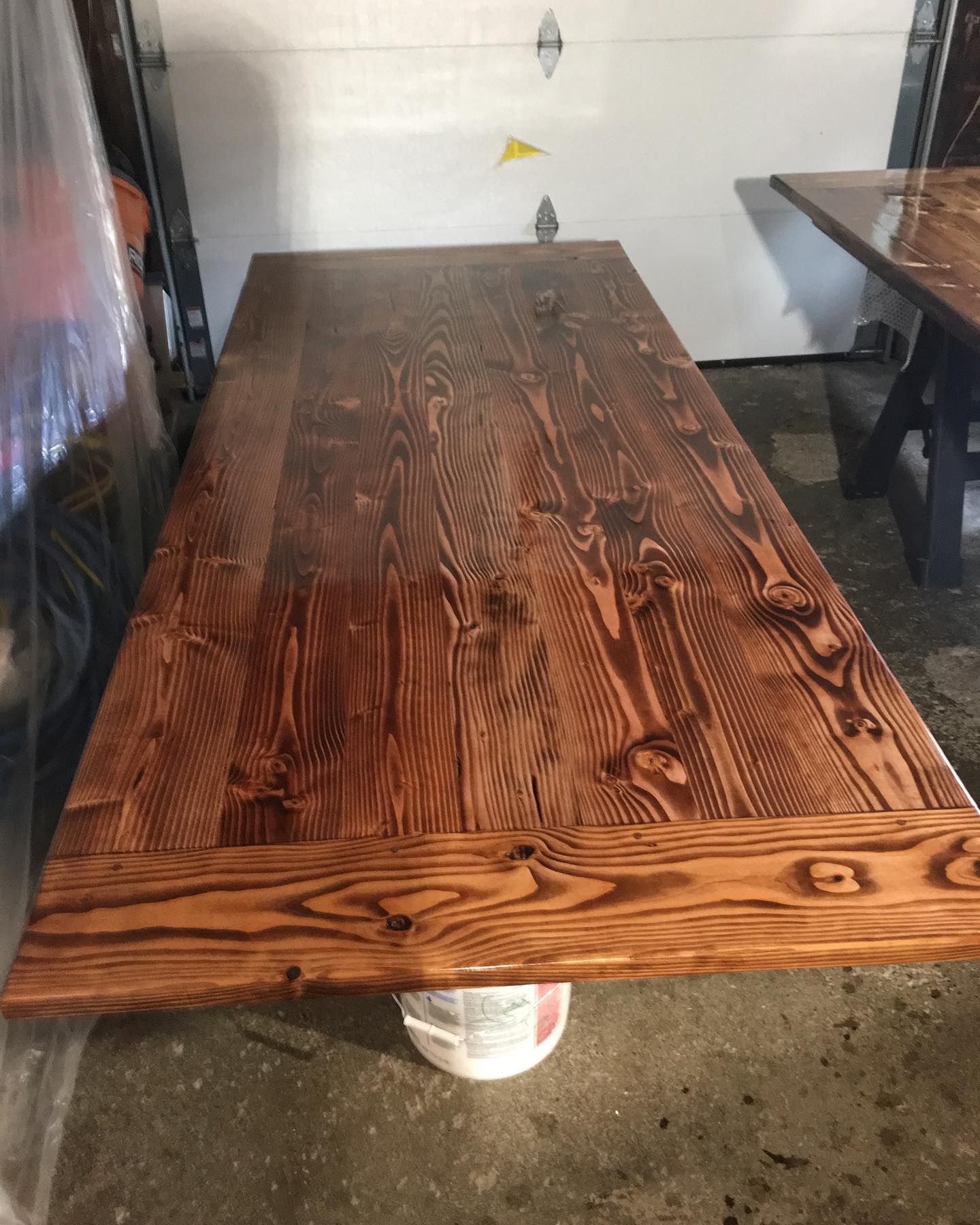 Wooden table top with stained wood grain, propped up on a white bucket in a garage.