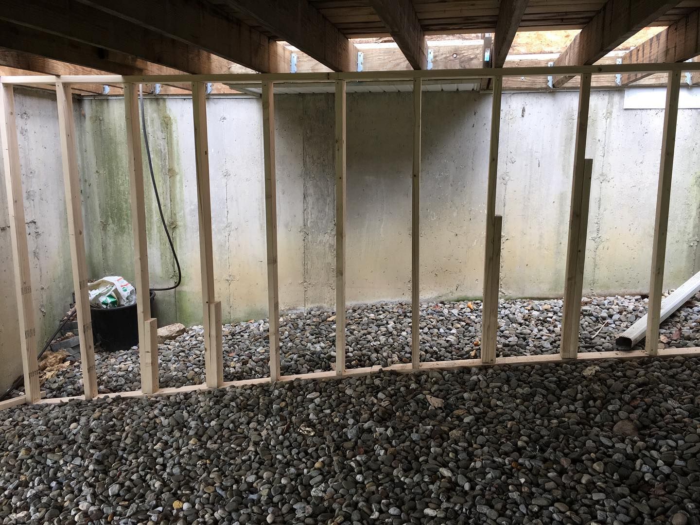 Framing for a basement wall. Wooden studs on a gravel floor against a concrete wall under wooden beams.