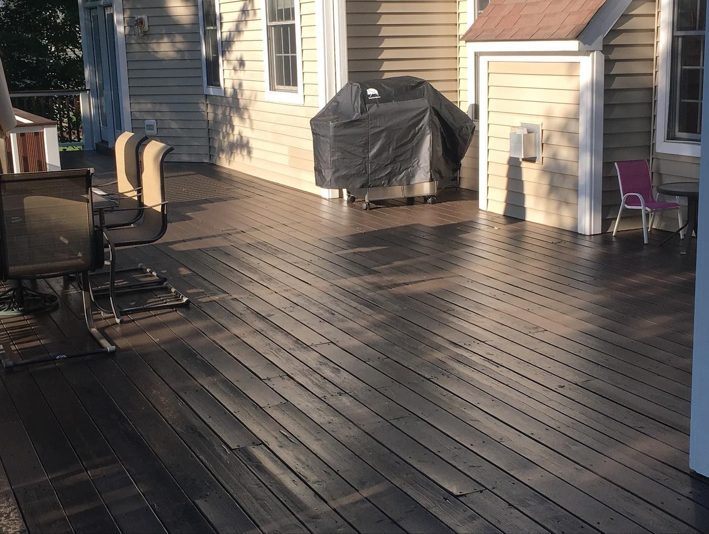 A wooden deck with grill, chairs, and the side of a house. Sunny day with light and shadow.