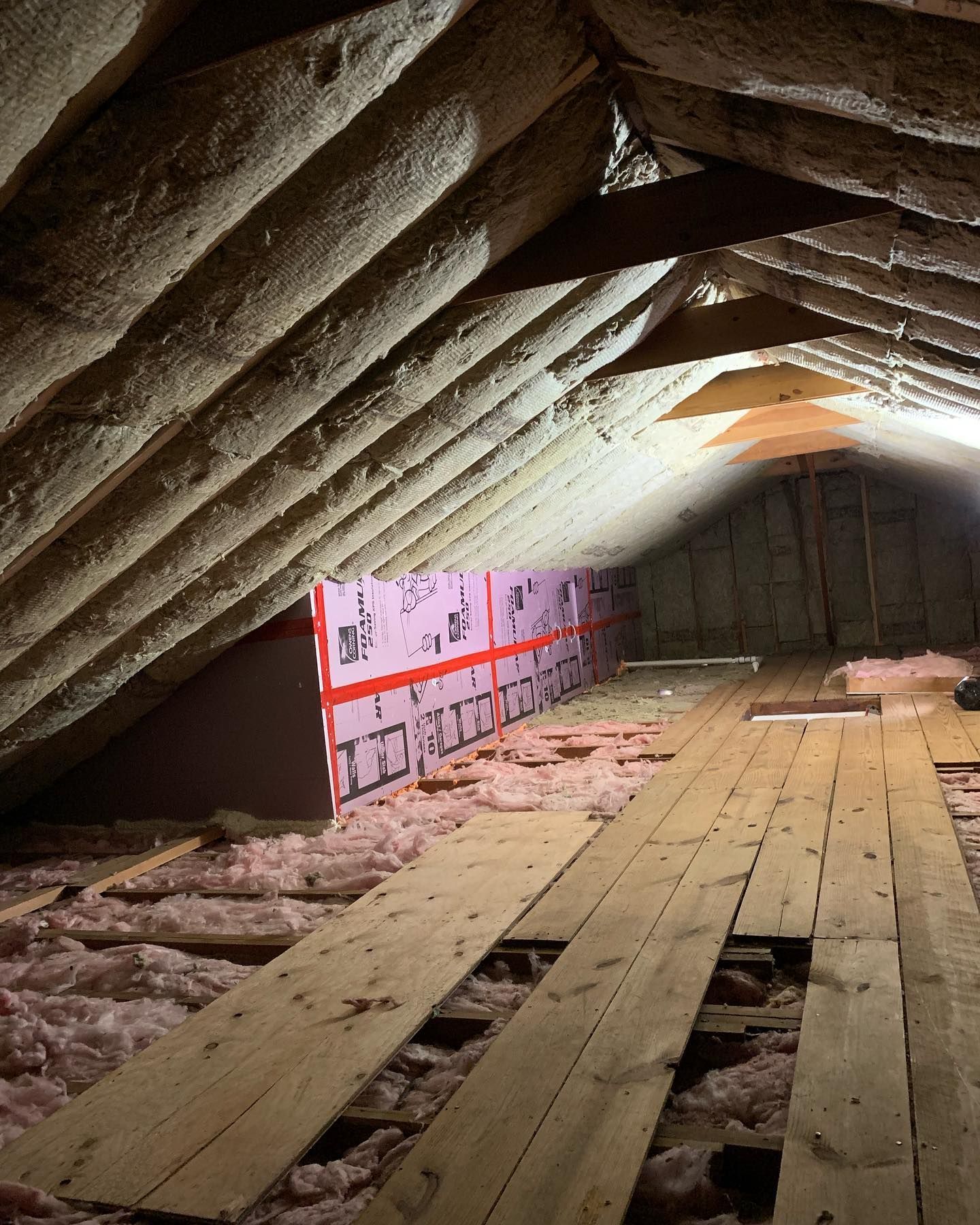 Attic space with wooden beams and planks. Pink insulation on a slanted wall, and pink fiberglass insulation on the floor.