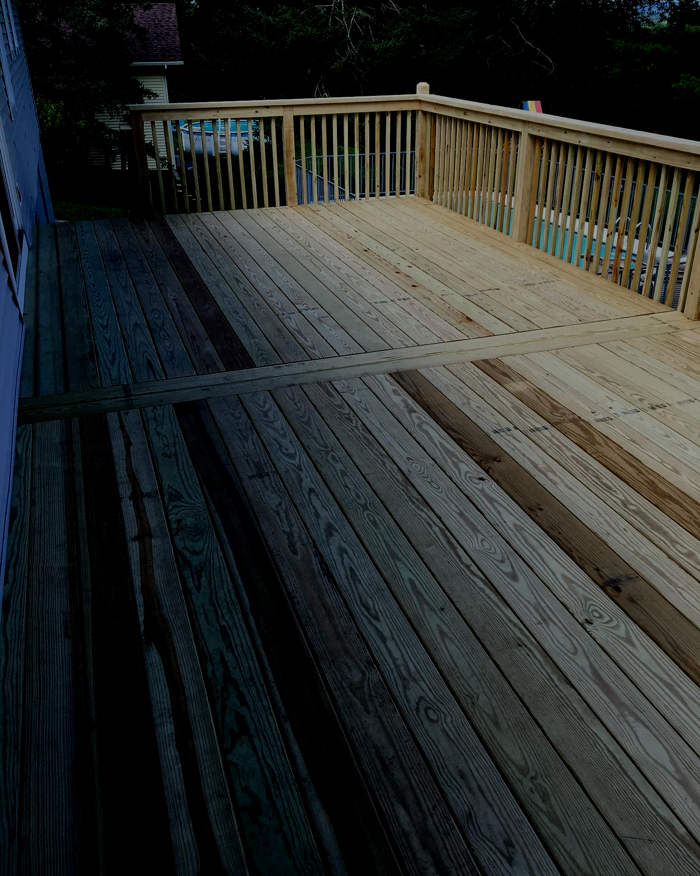 Wooden deck with a railing, connected to a house. Part of the deck is in shadow.