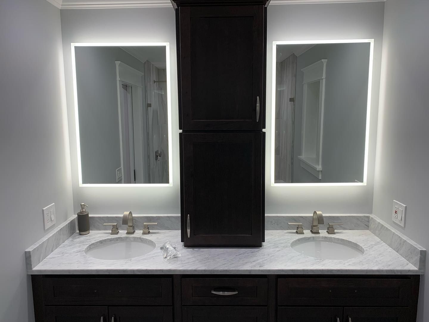 Bathroom with two illuminated mirrors, dark cabinets, and granite countertop.