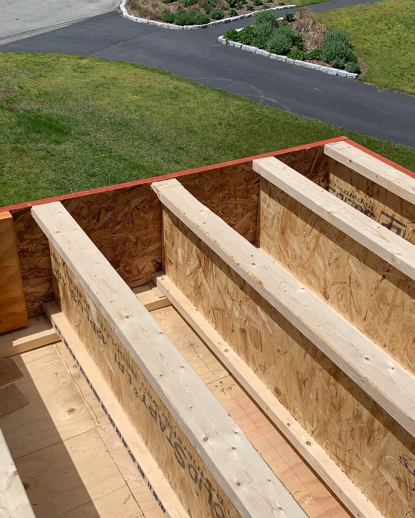 Wooden beams on a deck frame, constructed from OSB and lumber, outdoors with green grass and a driveway in the background.
