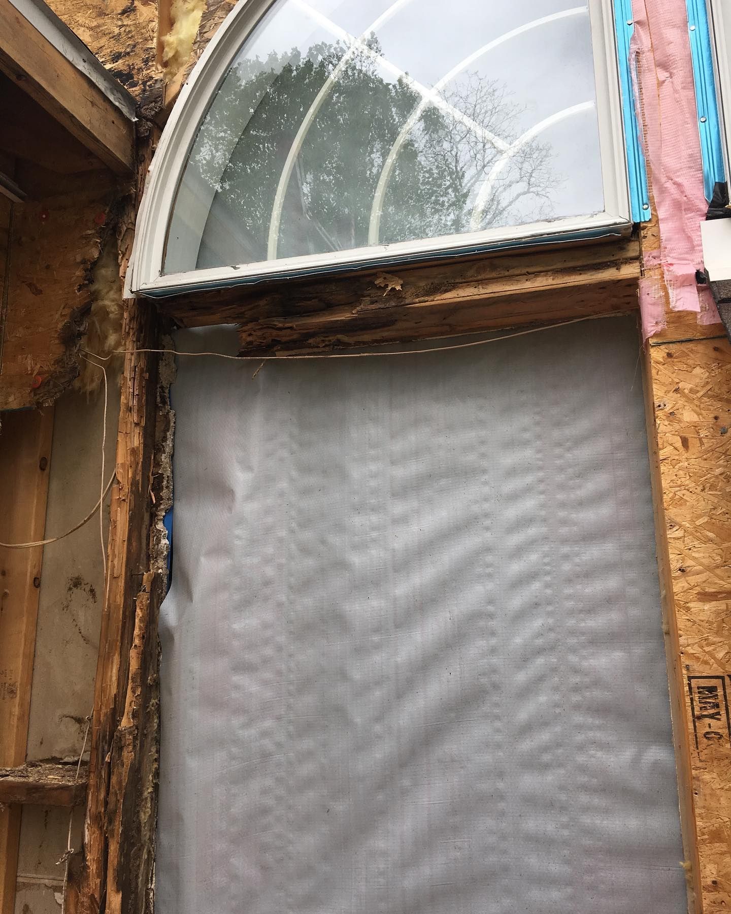Exterior view of a window with water damage and exposed framing, covered by a gray barrier.
