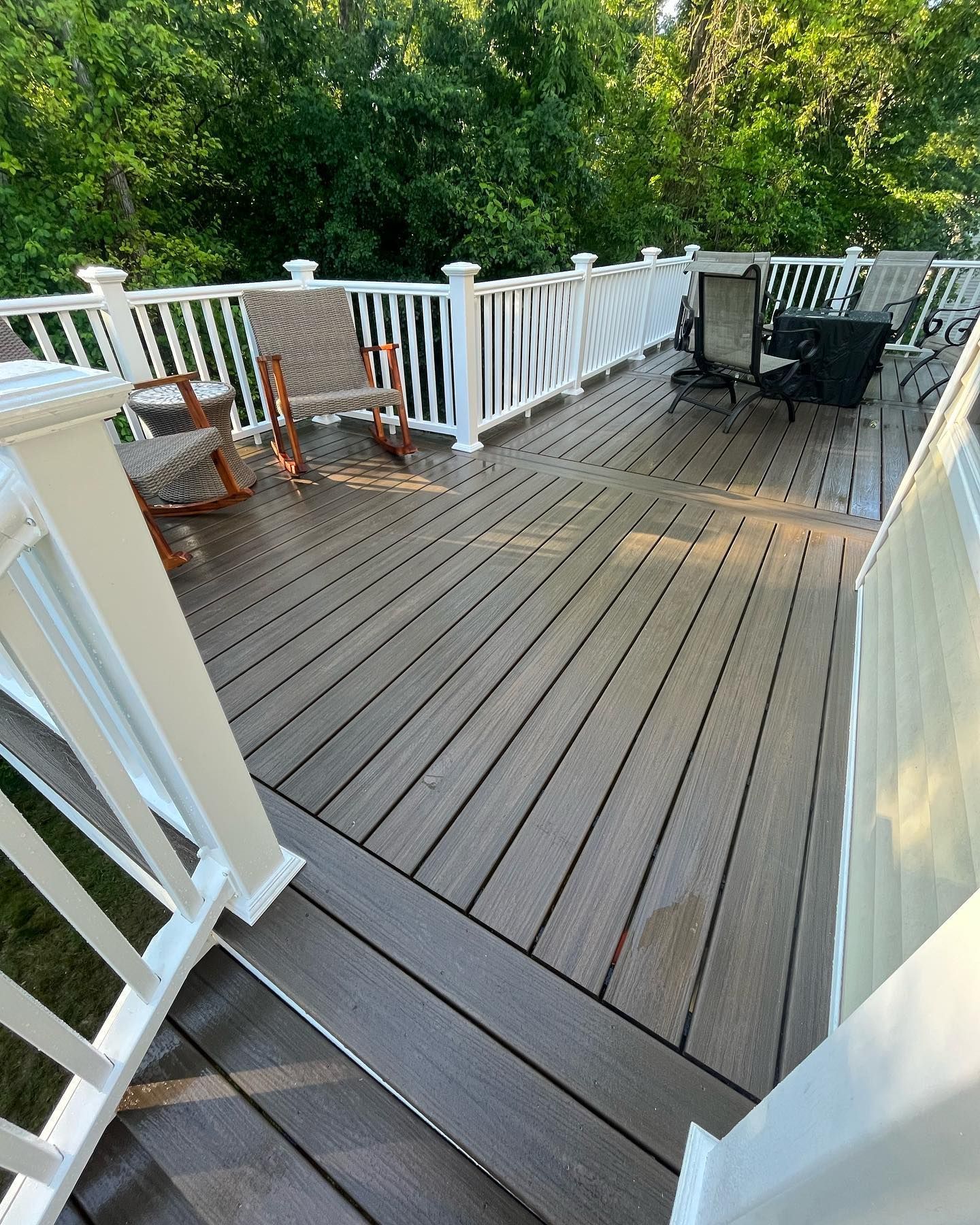 Wooden deck with white railing, two rocking chairs, and dark seating, surrounded by trees.