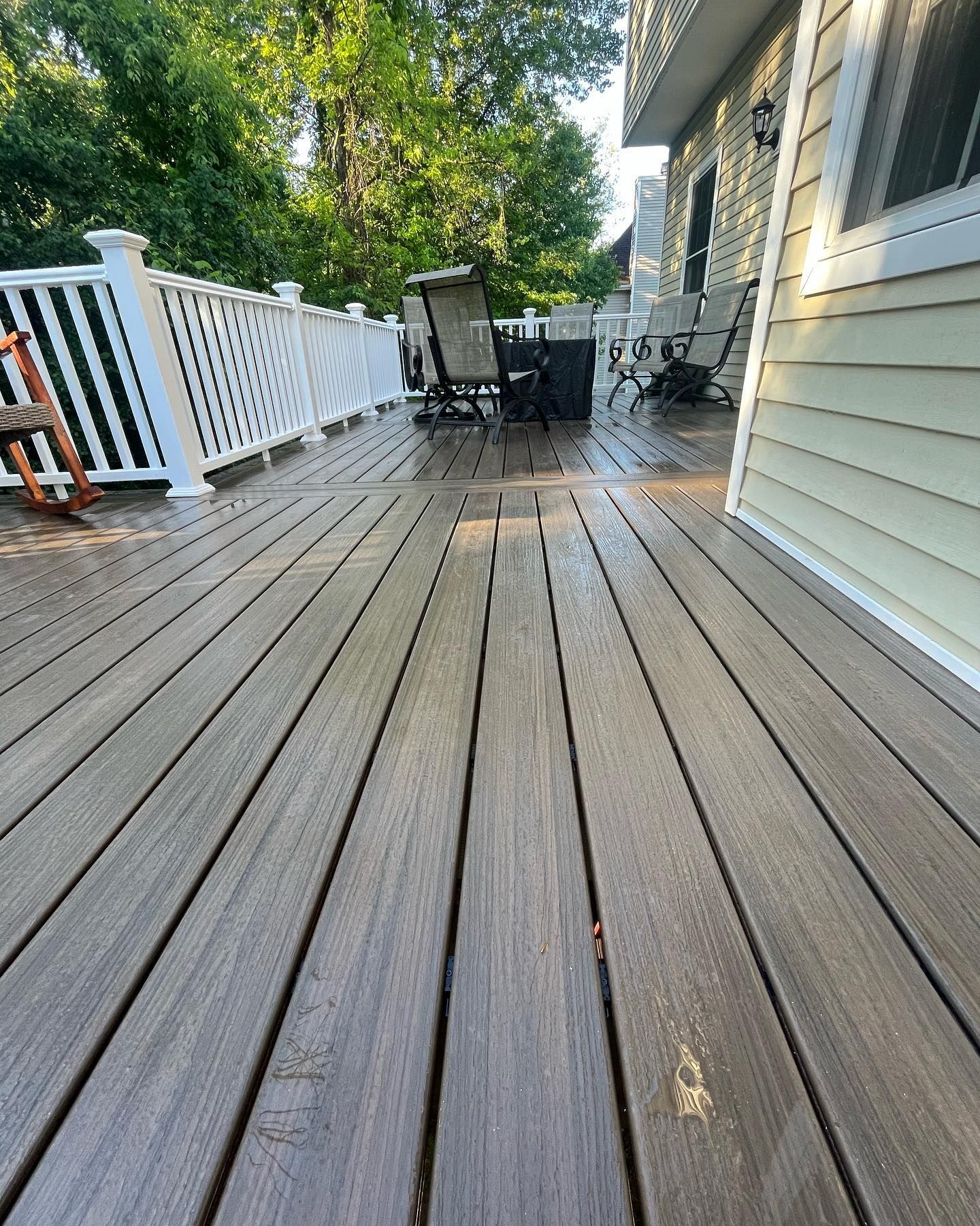 Wooden deck extending from a light yellow house, with chairs and trees in the background.