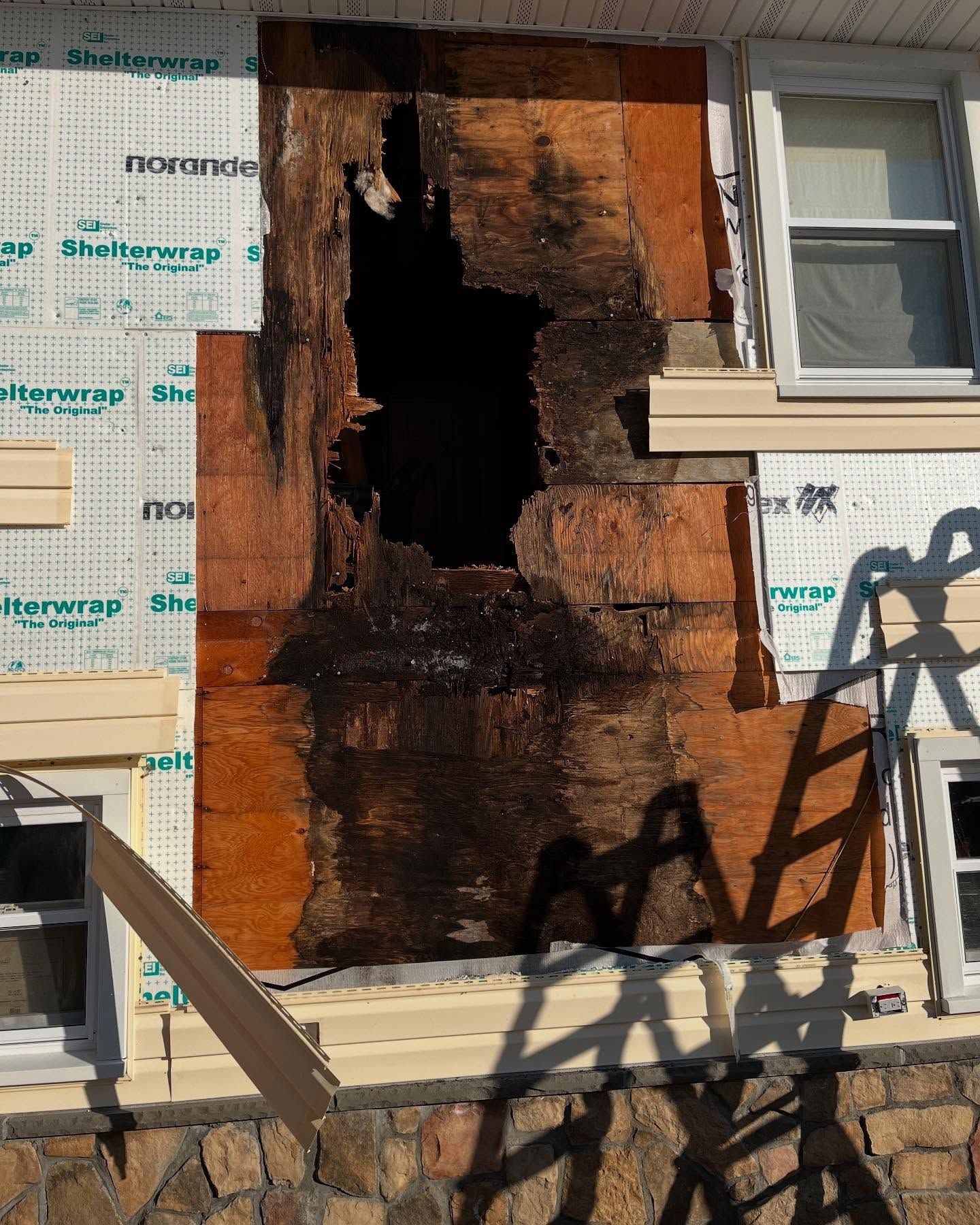 Damaged exterior wall with a large hole, water damage, covered in sheathing and adjacent to a window.