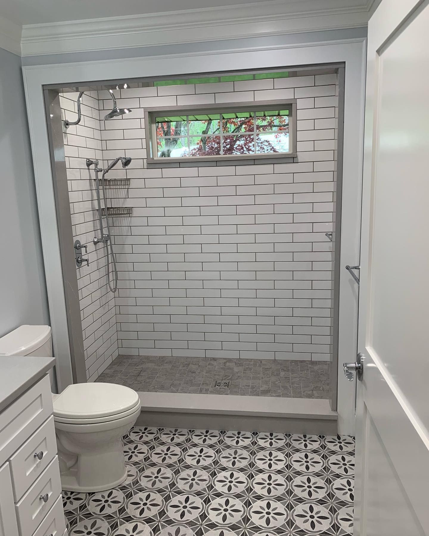 Bathroom with patterned floor, white brick-like shower, window, toilet, and white vanity.