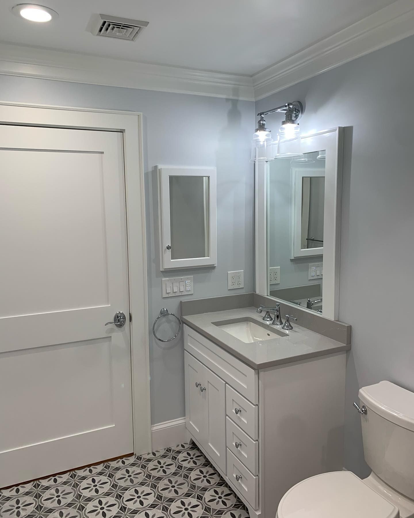 Bathroom with white vanity, mirror, and toilet; light blue walls, patterned tile floor, and a white door.