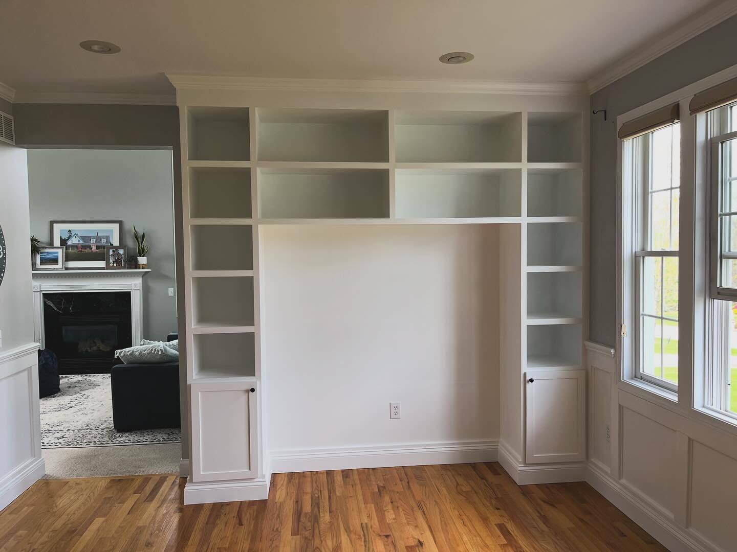 Built-in white bookshelf with open shelves and cabinets, in a room with wood floors and a window.