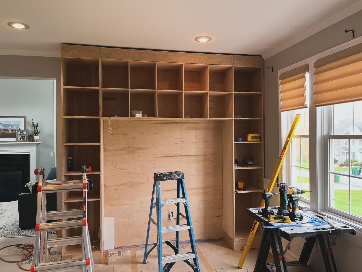 Wooden built-in bookcase under construction in a room with a window. A ladder and tools are present.