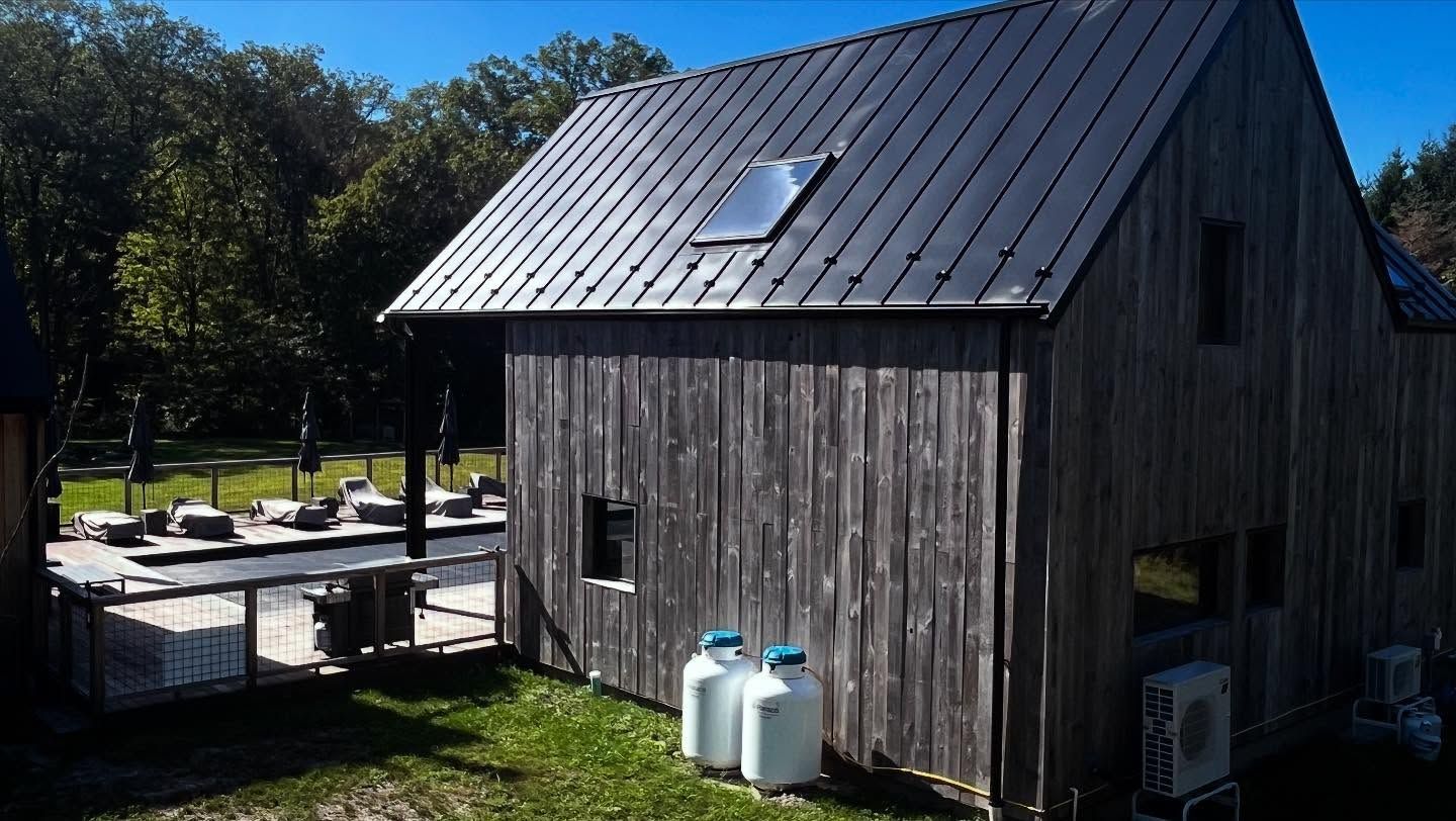 Wooden barn with dark metal roof, propane tanks, and a solar panel against a backdrop of trees and blue sky.