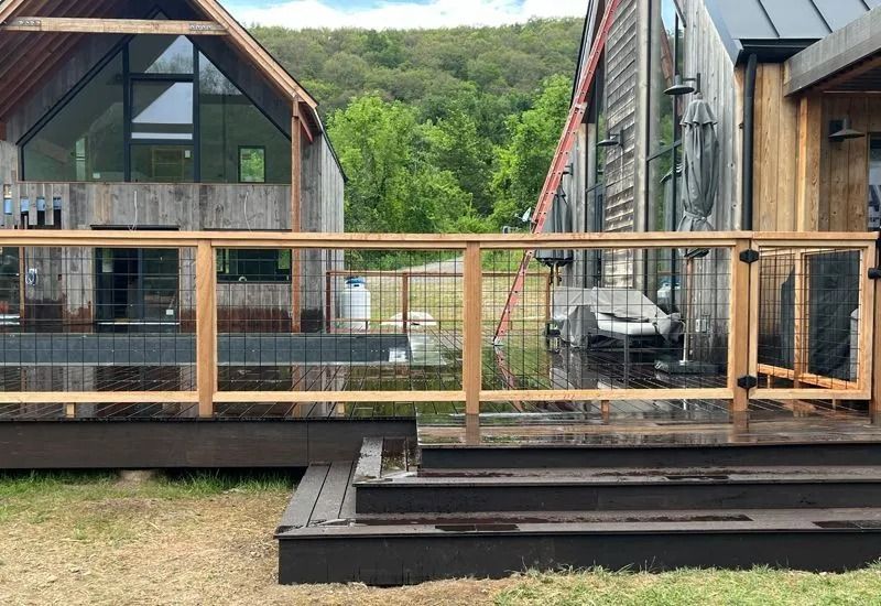 Wooden deck with wire mesh railing and steps, beside rustic buildings, green background.