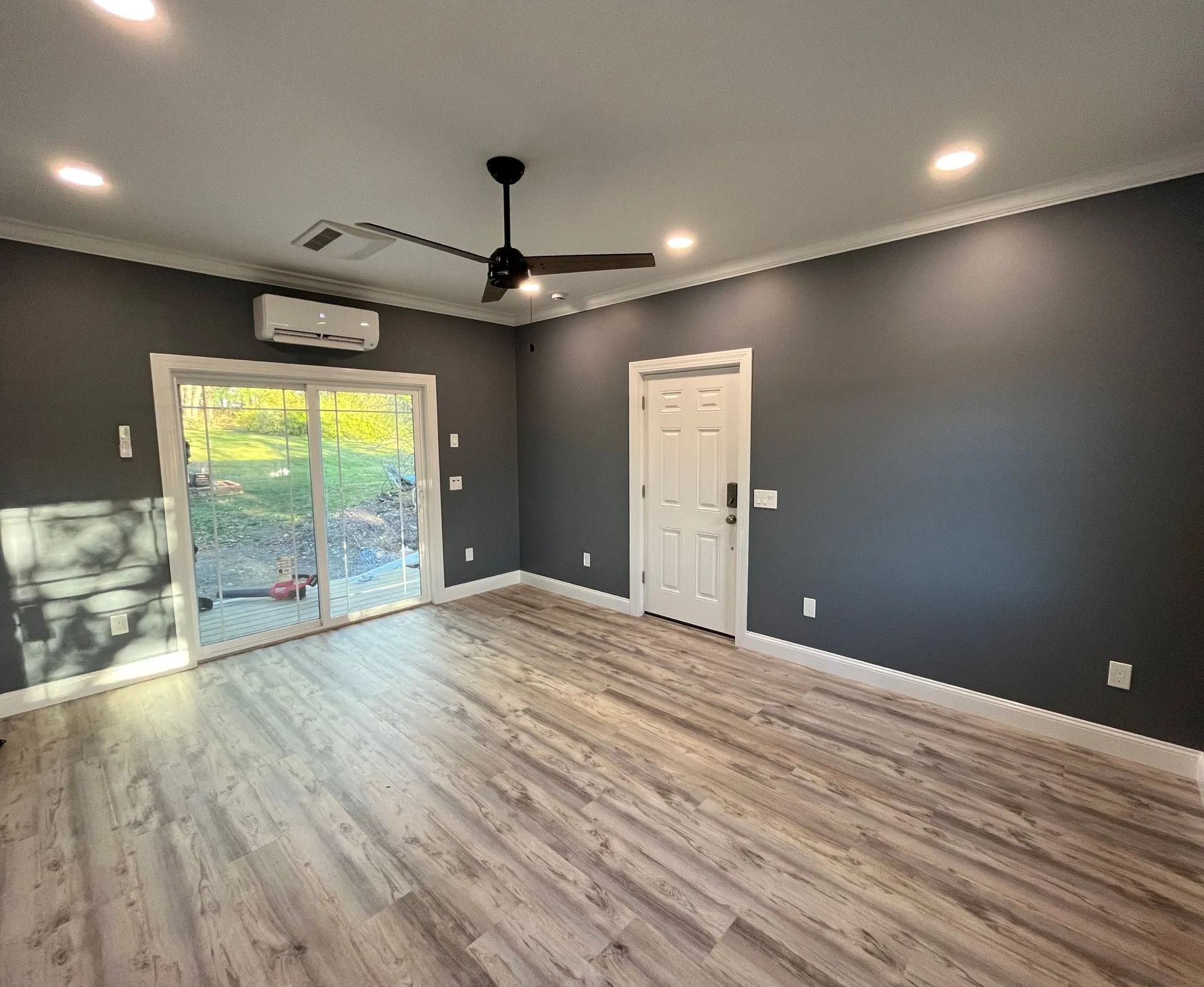 Empty room with gray walls, light wood floor, sliding glass door, and white door. A ceiling fan and AC unit are also visible.