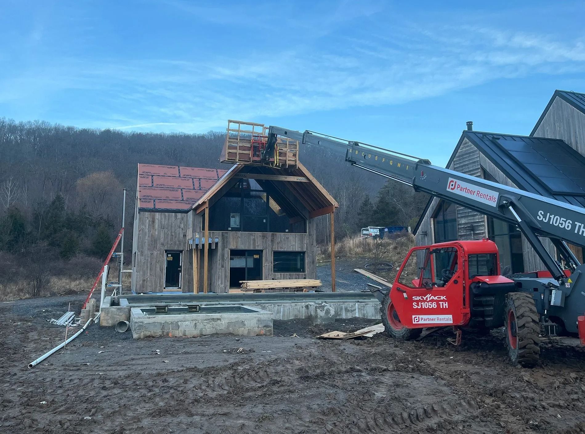 Construction site with a red forklift lifting lumber onto a partially built wood-framed house. Cloudy day.
