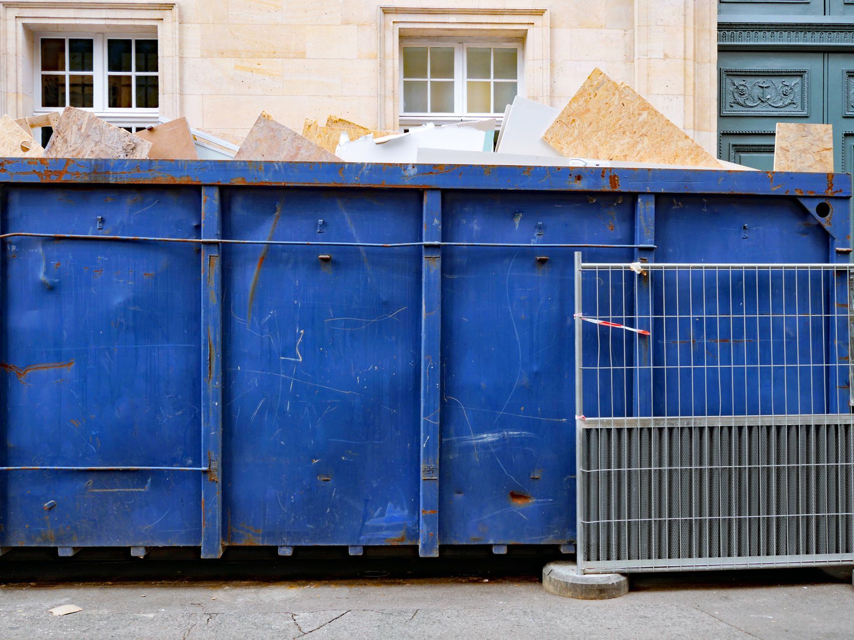 A blue dumpster is sitting in front of a building next to a fence.