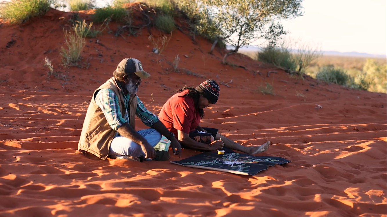 Two Indigenous people on red sand, making art outdoors.