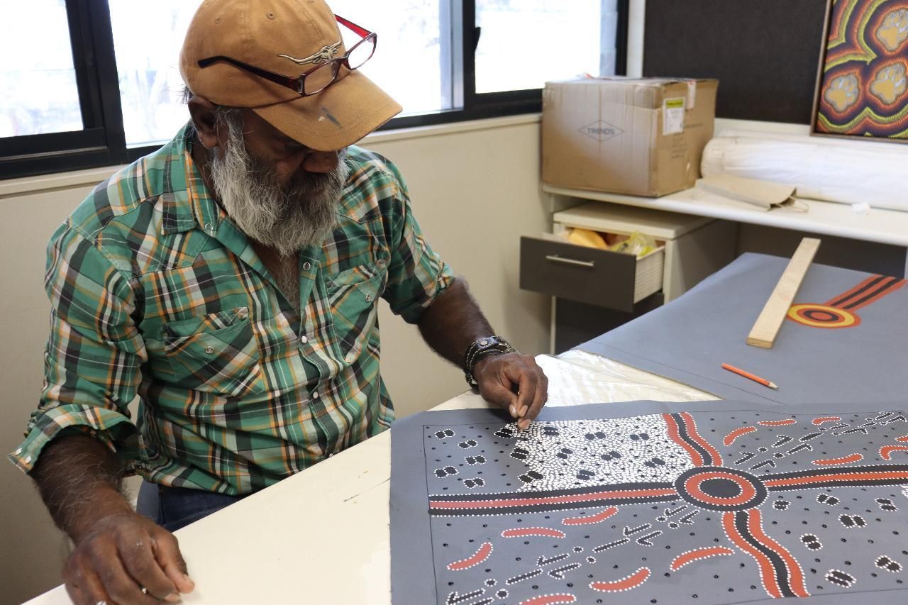 Man with a beard and hat carefully works on a colorful dot painting at a desk.