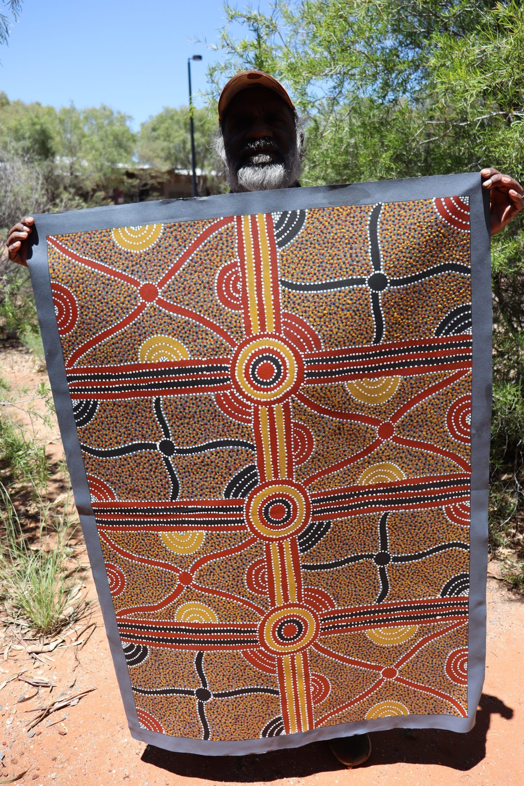 Man holding up a large, colorful Aboriginal artwork outdoors; browns, oranges, and blacks.